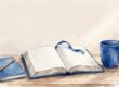 An open Bible, blue mug, journal, and pencil sit on a sunlit wooden desk.